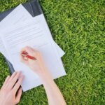 Руки підписують документи на чорній папці серед зеленої трави